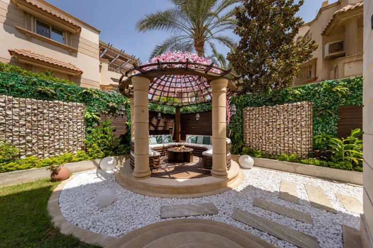 A landscape and outdoor living design by Aghbary Tex in Egypt. A round seating area with columns and a roof is placed in the center. A table and seating are set under the structure.