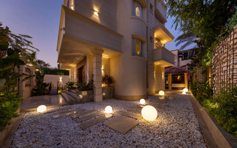 Exterior design by Aghbary Tex. A beige house with columns and balconies is featured. Round lights are set on a stone path. Plants and a stone wall are placed in the yard