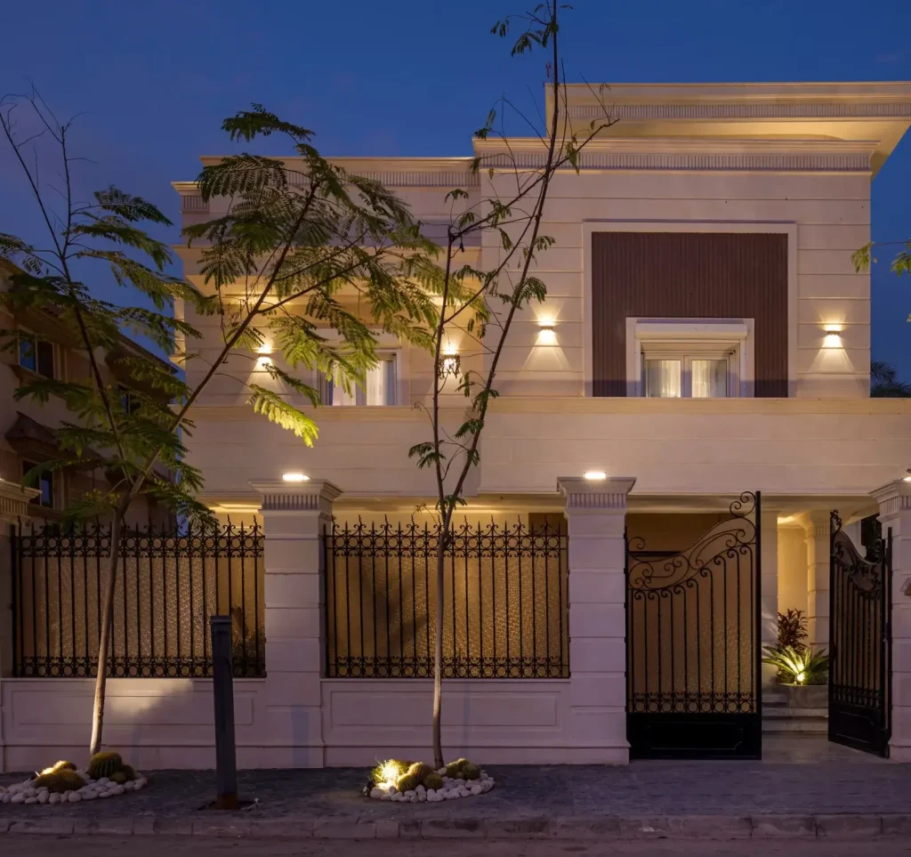 Exterior design by Aghbary Tex. A beige house with columns and wall lamps is featured. A black gate and fence are placed in front.